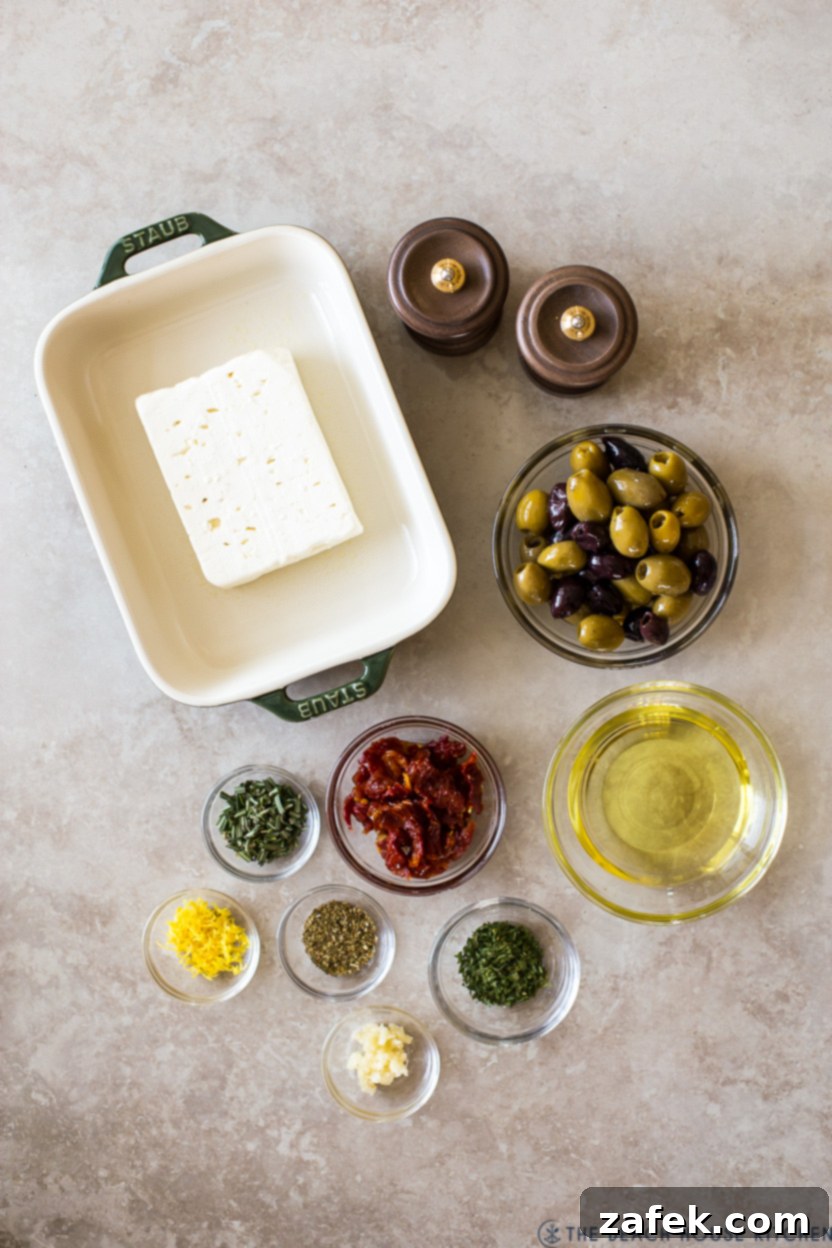 Overhead photo of the fresh ingredients artfully arranged for Baked Feta with Olives and Sun-dried Tomatoes, including a block of feta, mixed olives, sun-dried tomatoes, fresh rosemary, lemon, and garlic.