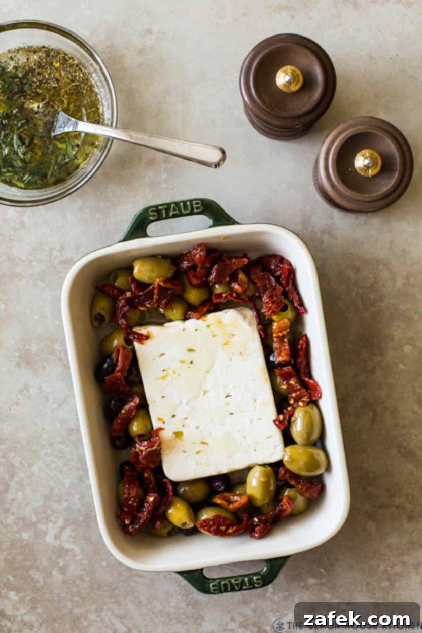 Overhead photo of the first step in preparing Baked Feta, showing a block of feta centered in a baking dish, surrounded by a colorful ring of mixed olives and sun-dried tomatoes.