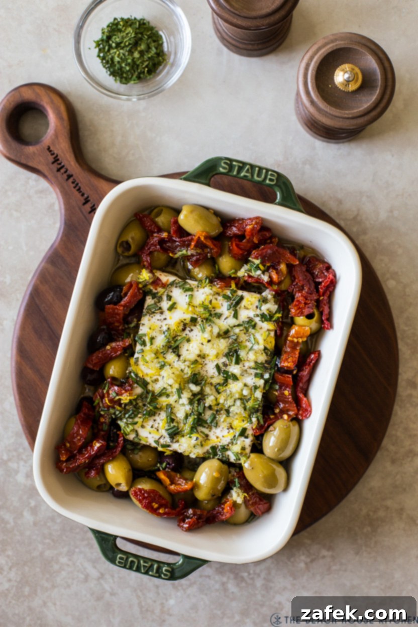Overhead photo of the second step in preparing Baked Feta, showing the block of feta and surrounding olives and sun-dried tomatoes generously drizzled with the prepared olive oil and herb mixture, ready for baking.