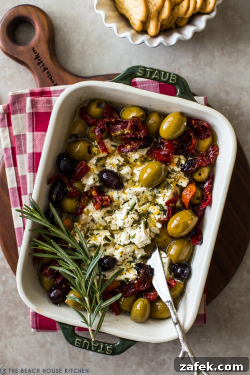 Up close overhead photo of the finished Baked Feta with Olives and Sun-dried Tomatoes, featuring the melted feta, vibrant olives, and sun-dried tomatoes, garnished with fresh herbs, and ready to be spread onto bread.