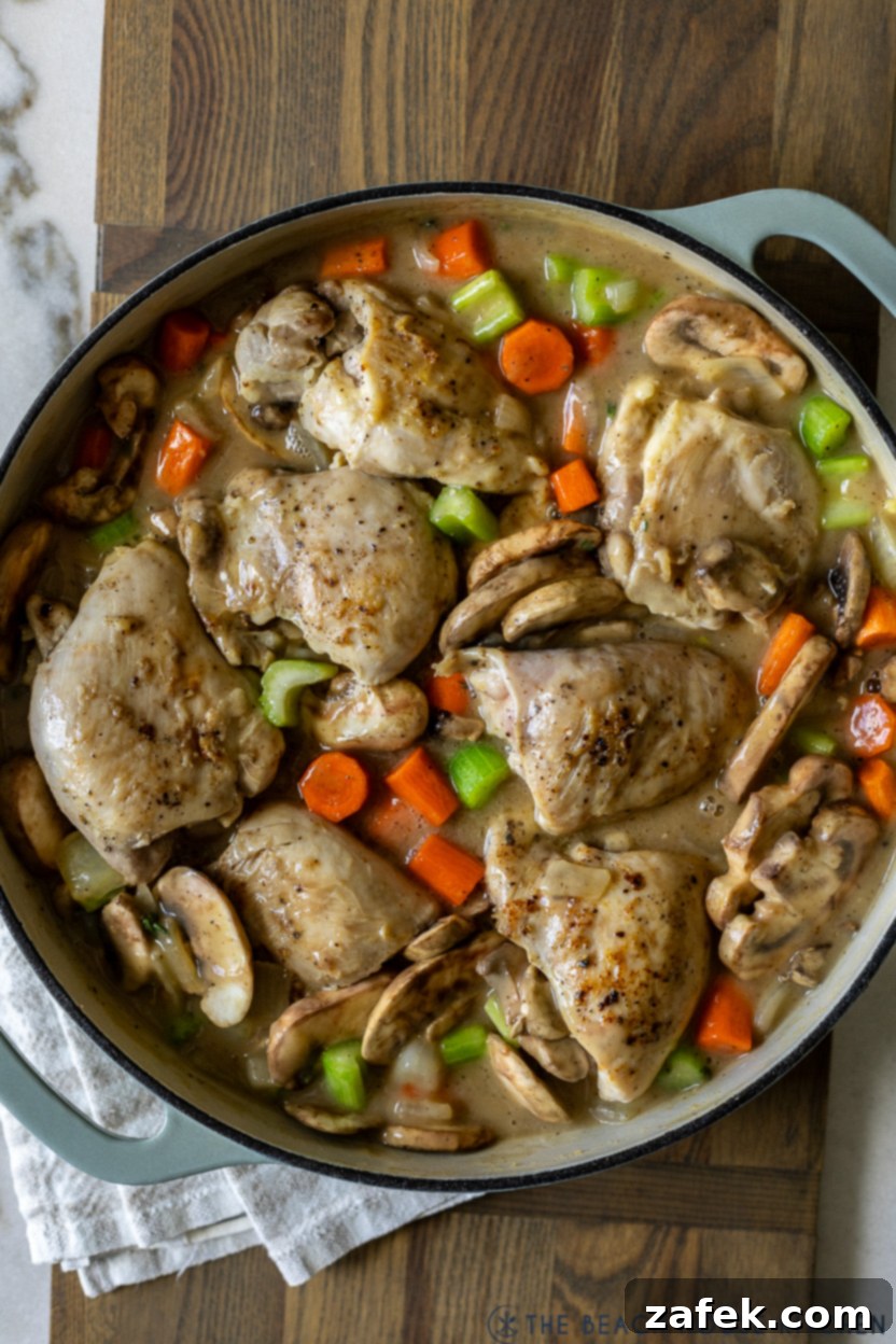Overhead photo of a skillet showing partially cooked chicken and mixed vegetables simmering in a light sauce, mid-cooking process for fricassee.