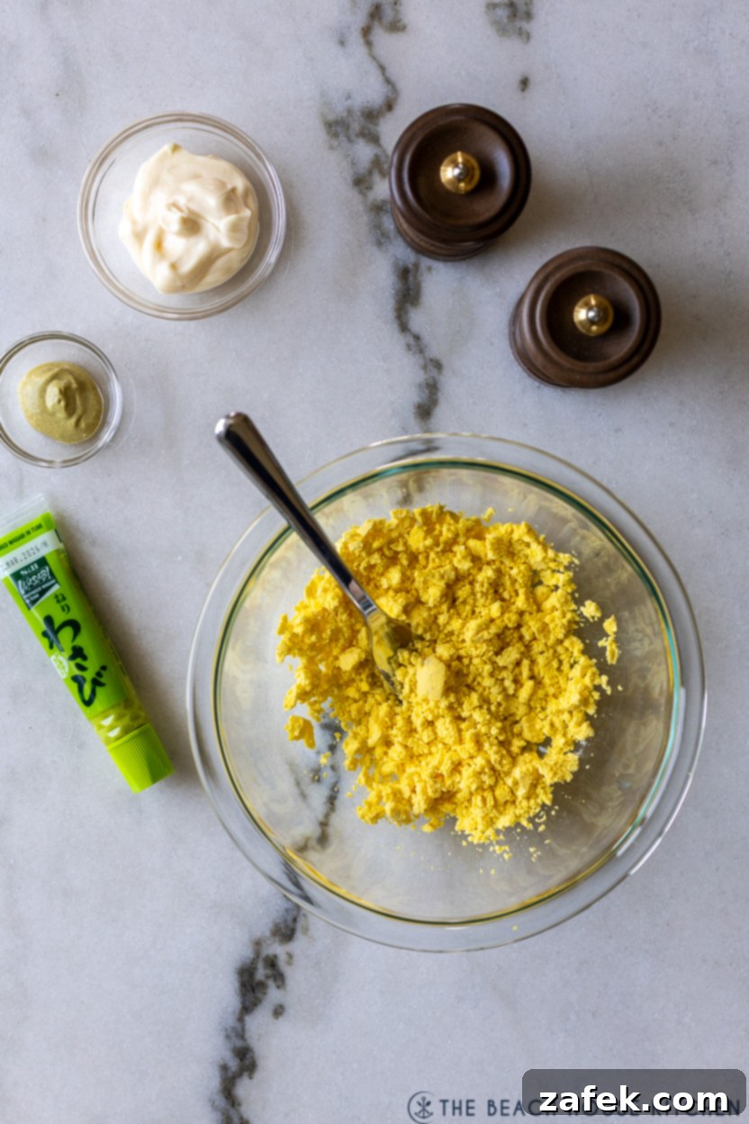 Overhead photo of a bowl of crumbled cooked egg yolks, a small bowl of mayonnaise, a dab of Dijon mustard, and a tube of wasabi paste, ready for mixing