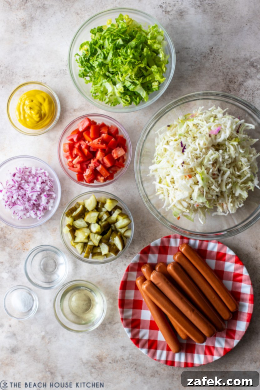 Overhead photo of fresh, colorful ingredients laid out for Chicago Hot Dog Salad preparation: neatly arranged sliced red onion, shredded cabbage, crisp romaine lettuce, diced ripe plum tomatoes, tangy garlic pickles, hot dogs ready for cooking, and a bowl with dressing components.