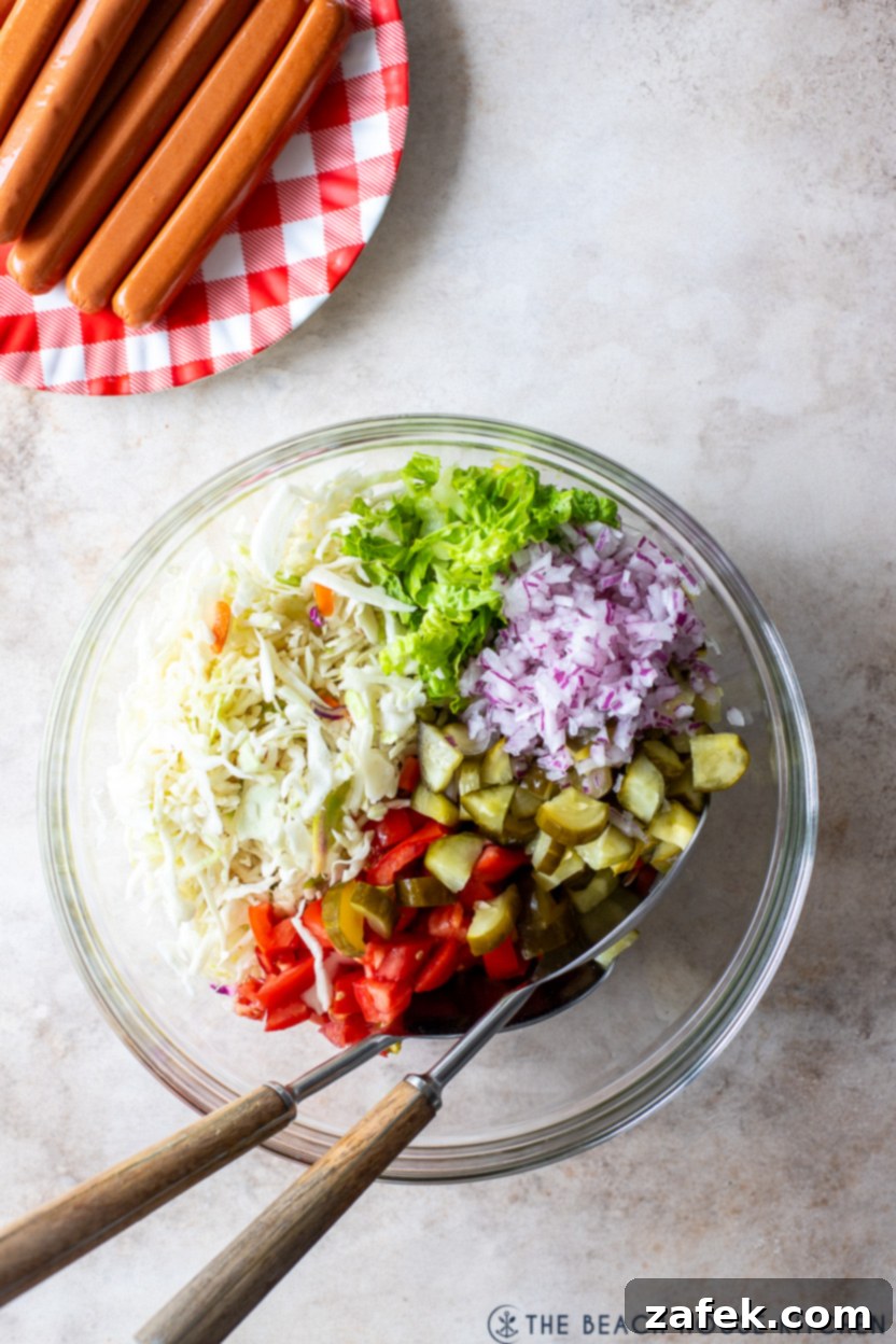 Overhead close-up photo of a large mixing bowl filled with vibrant fresh vegetables for Chicago Hot Dog Salad, including crisp lettuce, shredded cabbage, ripe tomatoes, red onions, and chopped pickles, all coated evenly in the zesty dressing.