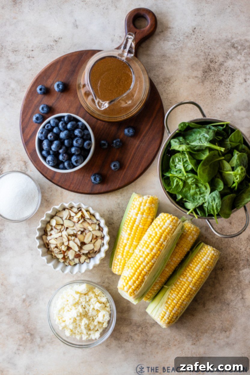 Overhead photo of a selection of fresh ingredients for blueberry corn salad, including corn on the cob, a pint of blueberries, spinach, almonds, and cotija cheese.