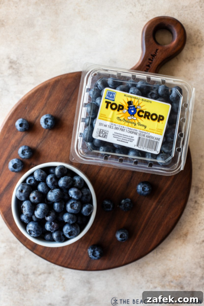 Overhead photo of a plastic container of fresh blueberries next to a small wooden bowl filled with blueberries, all resting on a round wooden cutting board.
