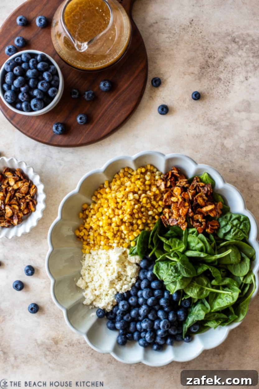 Overhead photo of a white serving dish meticulously arranged with individual piles of spinach leaves, blueberries, cooked corn, crumbled cotija cheese, and candied almonds, ready for tossing.