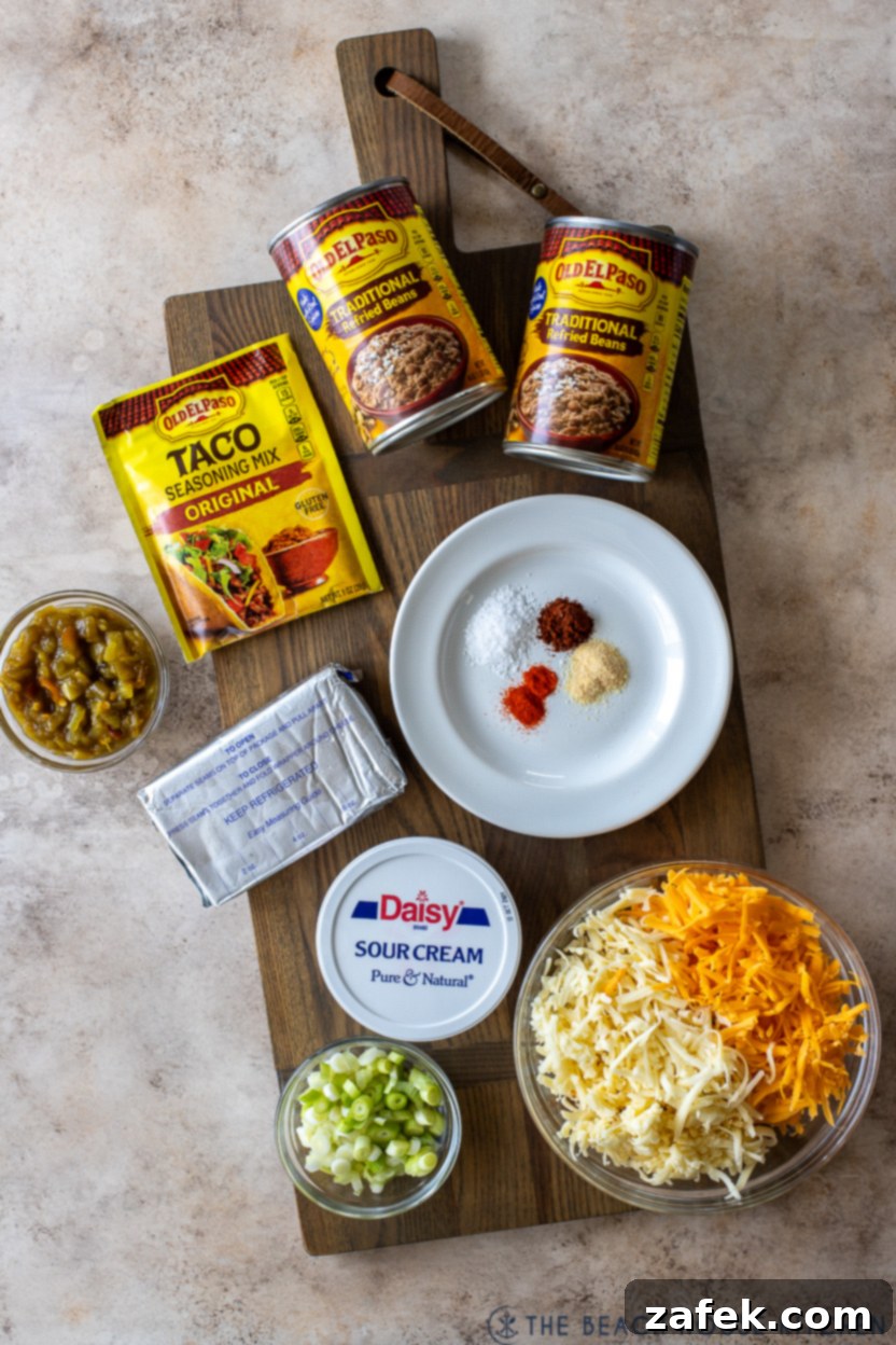 A vibrant overhead photo showcasing all the fresh ingredients for Texas Trash Dip laid out neatly on a kitchen counter, including cheeses, sour cream, refried beans, green chiles, and various spices.